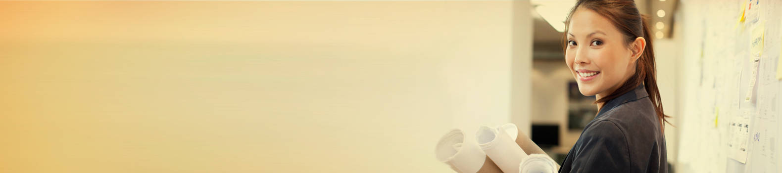 a lady holding blueprints, looking back and smiling, at corridor with notices on the wall