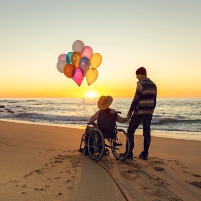 a disabled on a wheelchair and holding balloons with a man on the beach when sunset