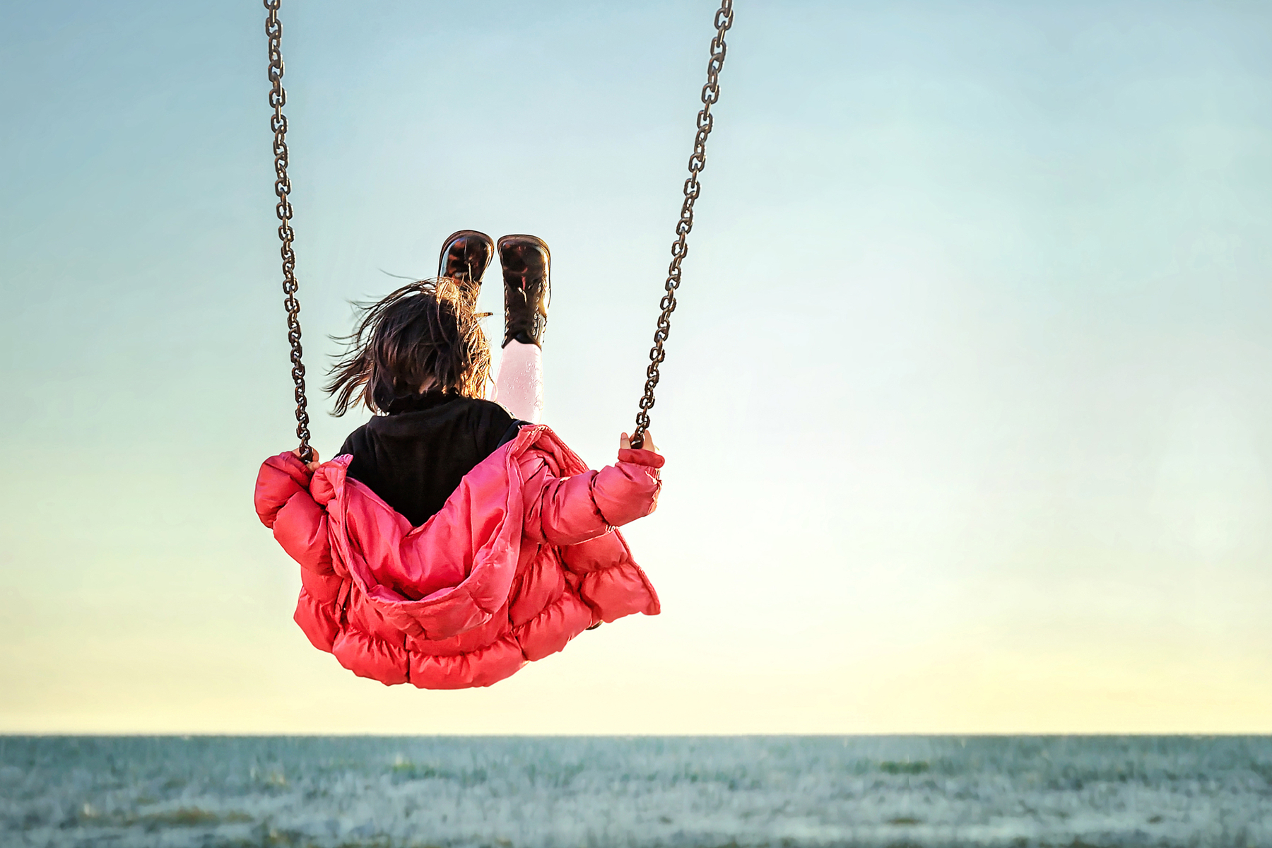 a little girl, wearing carol color down jacket, on a swing by the sea.